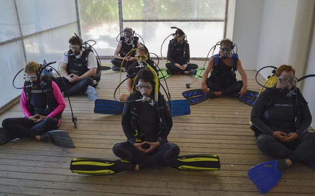 Image shows people in wetsuits and snorkeling gear meditating in a small room. 