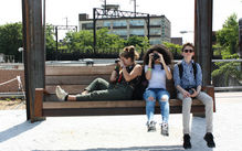 Three students are sitting on city park bench taking photos with digital cameras