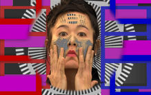 Artist Hito Steyerl in front of a brightly colored background comprised of magenta, blue, and black and white stripes.