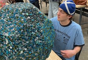 A student working on a large sculpture made out of blue and green beads. Other students work on similar shaped sculptures in the background, made of varying materials including playing cards and corks