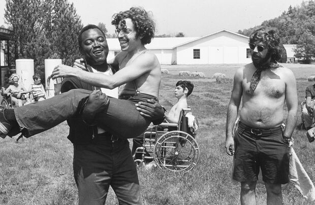 Black-and-white photograph of a joyful outdoor scene. A man carrying another man, both smiling, with people in the background, including individuals in wheelchairs, gathered in a field with buildings nearby.