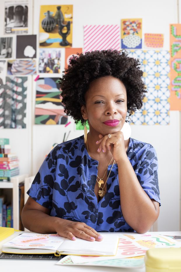 A woman with curly hair sits at a desk, looking confidently at the camera, with colorful artwork behind her.