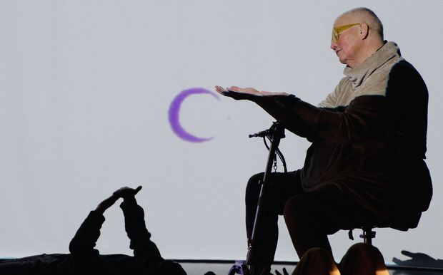 Petra,  a white queer disabled cis woman of size, sits on her scooter and reaches out her hand to a video projection of a purple moon. Below, a floor dancer reaches up and touches their own hands. Photo: Video still, Petra Kuppers 