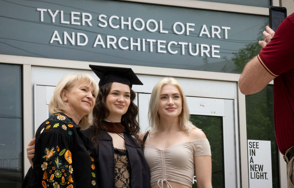 A graduate poses for a photo between two family members in front of the Tyler School of Art and Architecture entrance sign