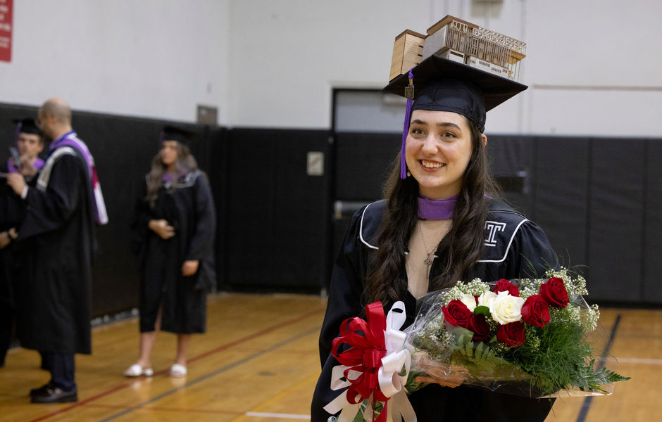 A graduate with a 3D architecture model on top of their mortarboard holding flowers 