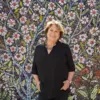 Sally Harrison smiles at the camera, standing in front of a vibrant mural filled with flowers.