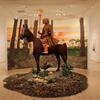Sculpted installation of a human rider on a horse atop a mound of earth. The installation is in front of a painted backdrop, but the white walls of the gallery are visible behind the life-size installation.