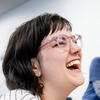 Close-up of a person laughing, wearing large light pink glasses and amber bead earrings.