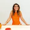 Portrait of Ali Ruffner wearing an orange dress leaning against a table, smiling at the camera.