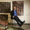 Gerard Brown sits on a metal stool with his legs extended straight out. Behind and to the left of him, large canvases featuring drawn artworks are displayed, creating an artistic and focused atmosphere around him.