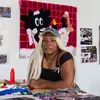 Portrait of Qualeasha Wood leaning against a studio table, with fiber artworks hanging in the background