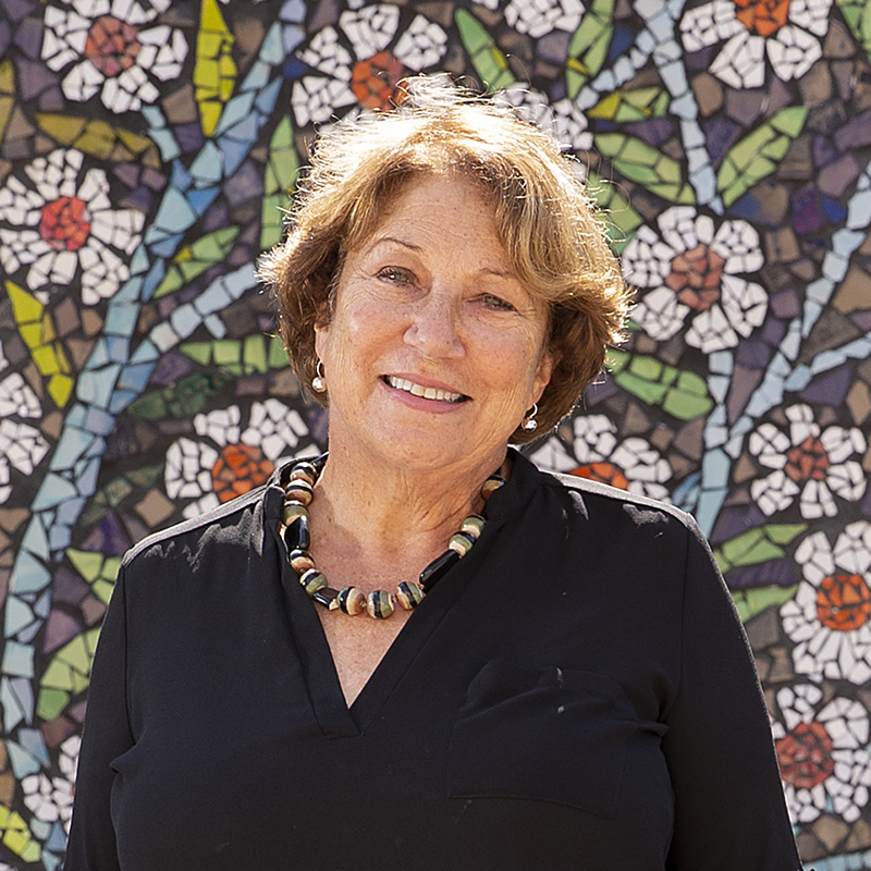Sally Harrison smiles at the camera, standing in front of a vibrant mural filled with flowers.
